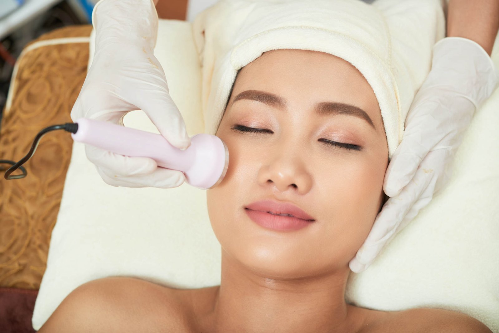 Close-up shot of beautician hands making ultrasound facial procedure while pretty Asian client lying on treatment table with closed eyes